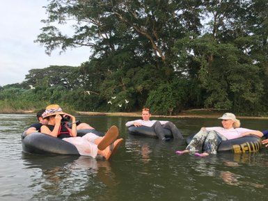 Fünf Personen entspannen auf Reifen im Wasser, umgeben von üppigem Grün und fliegenden Vögeln im Hintergrund.