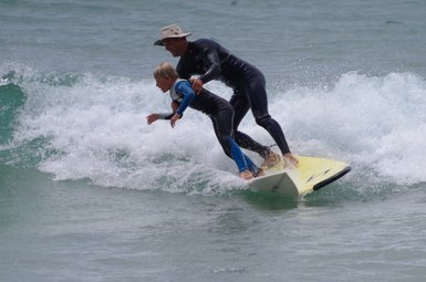 Ein Erwachsener und ein Kind surfen gemeinsam auf einer gelben Surfbrettwelle, umgeben von spritzendem Wasser.