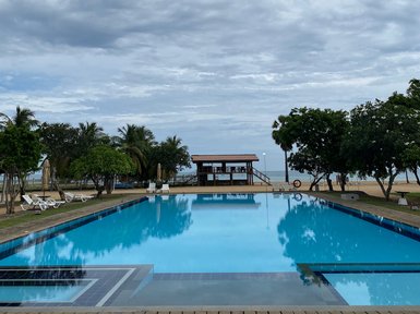 Ein weitläufiger Pool spiegelt den bewölkten Himmel wider, umgeben von Palmen und einer einladenden Liegewiese.