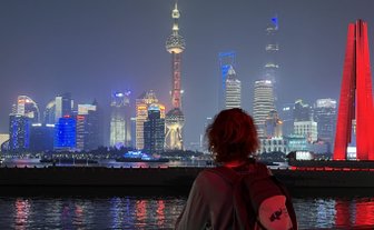 Eine Person mit lockigem Haar steht an einem Geländer und blickt auf die beleuchtete Skyline von Shanghai bei Nacht.