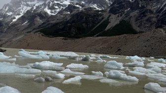 Im Vordergrund schwimmen Eisblöcke in einem trüben Gewässer, während majestätische Berge im Hintergrund aufragen.