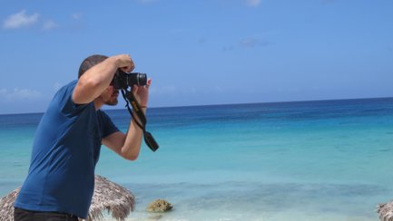 Ein Mann steht am Strand und fotografiert mit einer Kamera, während das türkisfarbene Wasser im Hintergrund leuchtet.