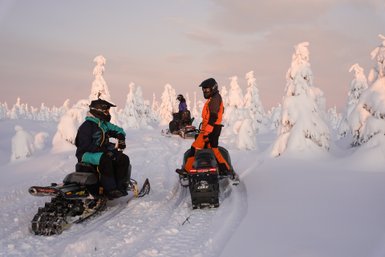 Zwei Schneemobilfahrer in winterlicher Landschaft, umgeben von schneebedeckten Bäumen und sanften Hügeln im Hintergrund.