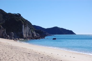 Ein ruhiger Strand mit feinem Sand, umgeben von steilen Klippen und klarem, blauem Wasser unter strahlend blauem Himmel.