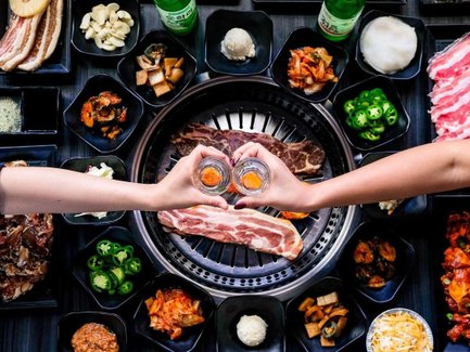 Zwei Hände halten Gläser mit einem orangefarbenen Getränk, umgeben von verschiedenen koreanischen BBQ-Gerichten auf einem Tisch.