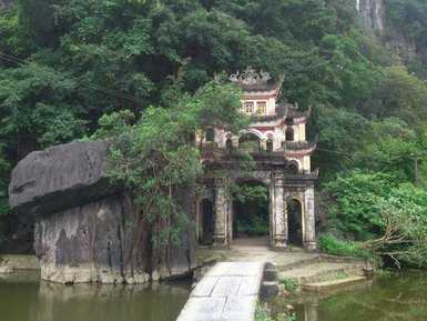 Ein malerischer Tempel mit bunten Verzierungen steht am Ufer eines ruhigen Teichs, umgeben von üppigem Grün und Felsen.