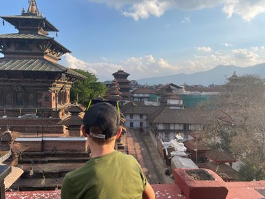 Ein Junge sitzt auf einem Dach und blickt auf die historische Architektur von Kathmandu, umgeben von Bergen und Wolken.