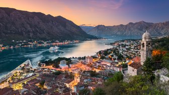 Ein atemberaubender Blick auf die Bucht von Kotor, umgeben von majestätischen Bergen und beleuchteten Städten bei Sonnenuntergang.