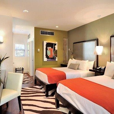 Ein modernes Hotelzimmer mit zwei Betten, orangefarbenen Bettdecken und einem stilvollen Schreibtisch in einem hellen Raum.