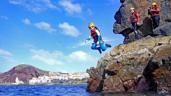Ein Junge springt fröhlich von einem Felsen ins Wasser, während andere Teilnehmer ihn beobachten und sich vorbereiten.