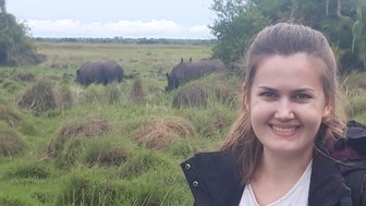 Eine lächelnde Frau steht in einer grünen Landschaft, während im Hintergrund zwei Nashörner grasen.