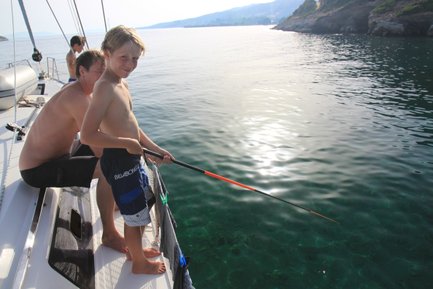 Ein Junge steht auf einem Boot und angelt, während ein Mann hinter ihm sitzt und die ruhige Wasseroberfläche betrachtet.