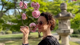 Eine Frau mit Sonnenbrille steht unter blühenden Kirschbäumen und hält einen Zweig mit rosa Blüten in der Hand.