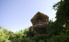 Eine Aussichtshütte aus Holz mit einem Strohdach, die auf einem großen Felsen inmitten üppiger Vegetation thront.