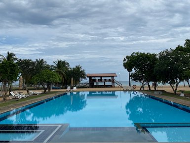 Ein weitläufiger Pool spiegelt den bewölkten Himmel wider, umgeben von Palmen und einer einladenden Liegewiese.