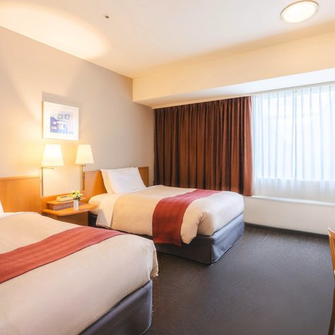 Ein helles Zimmer mit zwei Einzelbetten, warmen Farben und einem großen Fenster, das Tageslicht hereinlässt.