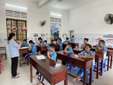 Eine Lehrerin steht vor einer Klasse, während die Schüler aufmerksam zuhören und in blauen T-Shirts gekleidet sind.