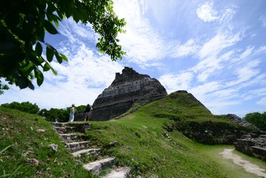 Zwei Personen stehen auf einer Wiese und betrachten die beeindruckenden Ruinen von Xunantunich unter einem strahlend blauen Himmel.
