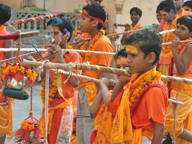 Eine Gruppe von Kindern in orangefarbener Kleidung, die mit Stöcken und Blumenkränzen an einem traditionellen Fest teilnehmen.