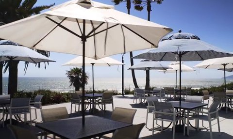 Ein sonniger Terrassenbereich mit weißen Sonnenschirmen und Tischen, der einen Blick auf den Ozean bietet.