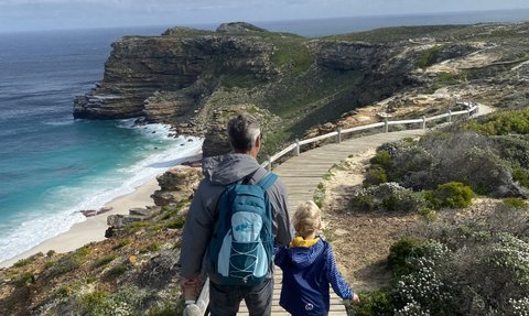 Ein Vater und sein Kind spazieren auf einem Holzsteg entlang der Küste, umgeben von üppiger Natur und dem Ozean.