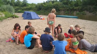 Eine Gruppe von Kindern sitzt im Sand am Flussufer, während sie sich um einen Jungen in Badebekleidung versammeln.