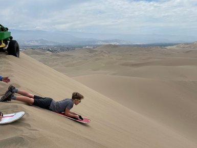 Ein Junge liegt auf einem Sandsurfbrett und bereit sich auf eine Abfahrt in die weiten Sanddünen vor ihm vor.