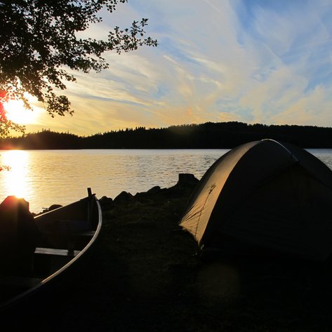 Ein Zelt steht am Ufer eines ruhigen Sees, während die Sonne hinter den Bäumen untergeht und den Himmel in warmen Farben färbt.