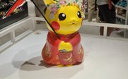 Ein Pikachu in traditioneller Kleidung mit einem roten Regenschirm steht in einem Geschäft voller Spielzeuge und Figuren.
