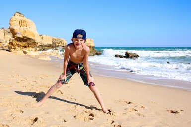 Ein fröhlicher Junge steht am Strand, mit weit gespreizten Beinen und einem breiten Lächeln, während die Wellen sanft an den Strand rollen.
