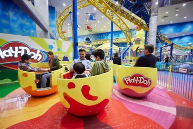 In einem bunten Indoor-Spielplatz sitzen Kinder in runden Fahrgeschäften, während Erwachsene in der Nähe zuschauen und lächeln.