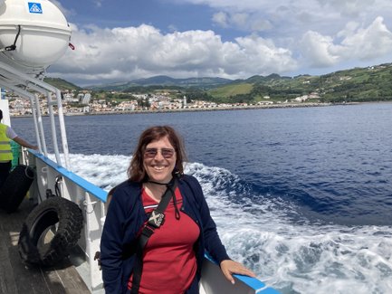 Eine lächelnde Frau steht auf einem Boot, während die Wellen hinter ihr spritzen und eine grüne Küstenlandschaft sichtbar ist.