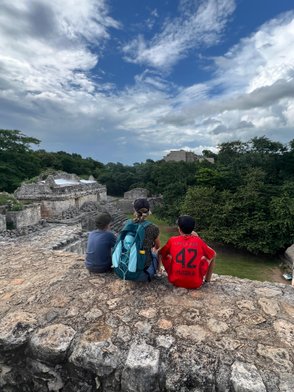 Drei Kinder sitzen auf einer alten Steinmauer und blicken auf die Ruinen einer historischen Stätte unter einem bewölkten Himmel.