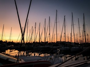 Ein malerischer Sonnenuntergang über einem Hafen, mit vielen Segelmasten, die sich gegen den bunten Himmel abheben.