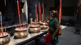 Eine Frau in einem grünen Kleid zündet Räucherstäbchen in einem Tempel an, während ein Kind neugierig zuschaut.