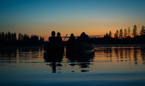 Vier Personen sitzen in Kanus auf einem ruhigen See, während die Sonne hinter den Bäumen untergeht und den Himmel färbt.