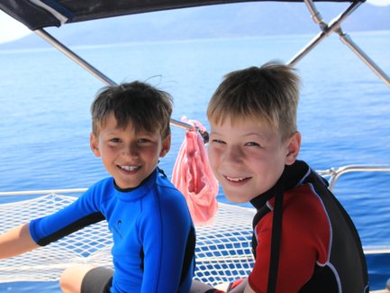 Zwei fröhliche Jungen in Neoprenanzügen sitzen auf einem Boot und genießen die Sonne und das Wasser um sie herum.