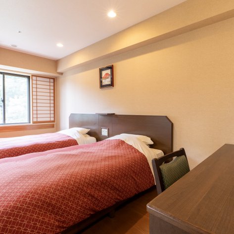 Zwei Einzelbetten mit roten Bettdecken stehen in einem hellen, modernen Zimmer mit einem großen Fenster.