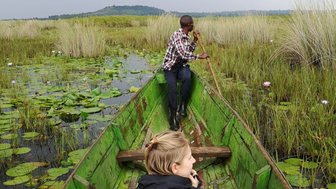 Eine Person paddelt in einem grünen Boot durch eine mit Seerosen bedeckte Wasserlandschaft, während eine andere Person zuschaut.