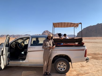 Ein Mann steht neben einem weißen Pickup, während zwei Kinder auf der Ladefläche sitzen und die Wüstenlandschaft betrachten.