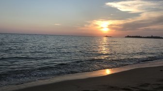 Ein malerischer Sonnenuntergang über dem ruhigen Wasser, das sanft den Sandstrand berührt und goldene Reflexionen erzeugt.