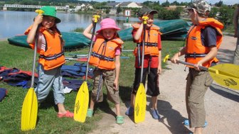 Vier Kinder in Schwimmwesten stehen am Ufer eines Gewässers, bereit zum Paddeln mit gelben Paddeln in der Hand.