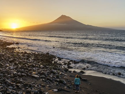 Eine Person steht am Strand und betrachtet den Sonnenuntergang über dem Meer, während der Vulkan im Hintergrund sichtbar ist.