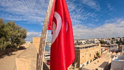 Die rot-weiße Flagge Tunesiens weht stolz über einer historischen Stadtansicht mit weißen Gebäuden und blauem Himmel.