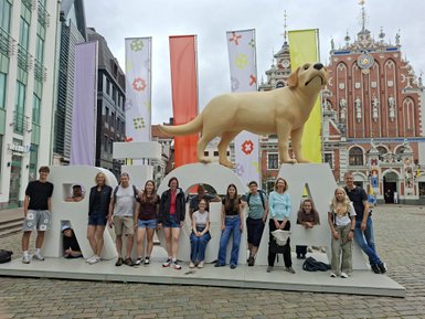 Eine Gruppe von Menschen posiert fröhlich vor einem großen, gelben Hund und bunten Fahnen in einer Stadt.