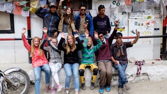 Eine Gruppe von Jugendlichen und Erwachsenen posiert fröhlich vor einer Wand mit bunten Gebetsfahnen in einem Dorf.