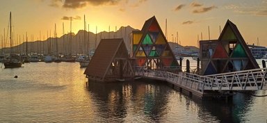 Bunte, pyramidenförmige Gebäude stehen am wasserumgebenen Kai, während die Sonne hinter den Bergen untergeht.