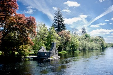 Ein ruhiger Fluss fließt sanft vorbei an einer alten Steinruine, umgeben von üppigem, grünem Laub und bunten Bäumen.