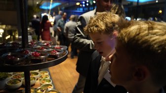 Zwei Jungen betrachten neugierig eine Auswahl an Desserts in Gläsern, während eine festliche Atmosphäre im Hintergrund herrscht.
