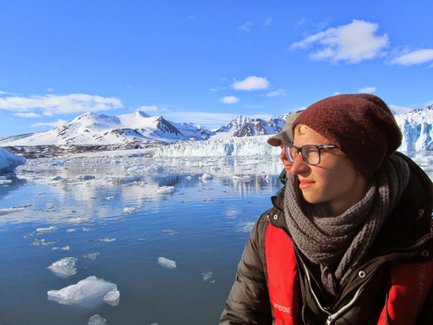 Eine Person mit Brille und Mütze blickt nachdenklich auf das ruhige Wasser, umgeben von schneebedeckten Bergen.
